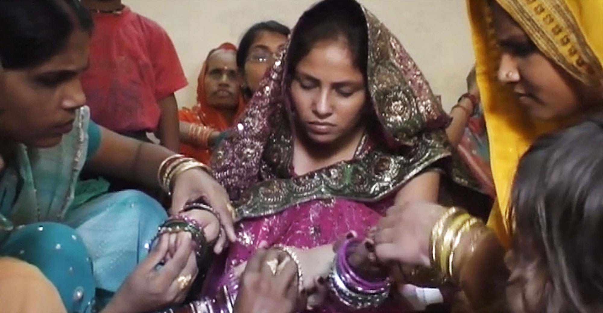 Кадр з фільму "Рішення". Середній план. Барвистий весільний індійський одяг. На передньому плані двоє жінок зосереджено вдягають прикраси на руки третьої, її обличчя напружене. Позаду сидять люди.