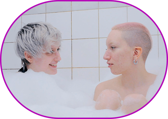 Still from the film "Always Amber". Two shot. Clean white tiles, two young people with piercing and colorful hairstyles sit in a foamy bathtub, looking at each other with love.