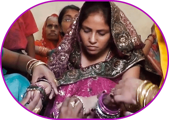 Still from the film "Decision". Medium long shot. Colorful Indian wedding clothes. Two women in the foreground carefully decorate the bride’s hands with jewelry, her facial expression is tense. There are people sitting in the background.