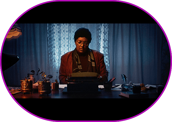 Still from the film "Octavia's Visions". Medium long shot. Blue-gray interior, warm light of a table lamp. A person wearing a jacket, scarf and glasses in the center of the image intently works on a typewriter.