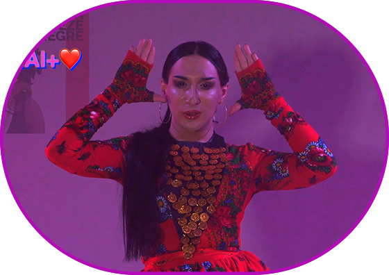 Still from the film "The Black Chick". Medium shot. A person wearing a bright red dress and golden jewelry raised the arms in the dance movement on the violet background, looking directly at the viewers. LGBTQAI+ is written on the left side.