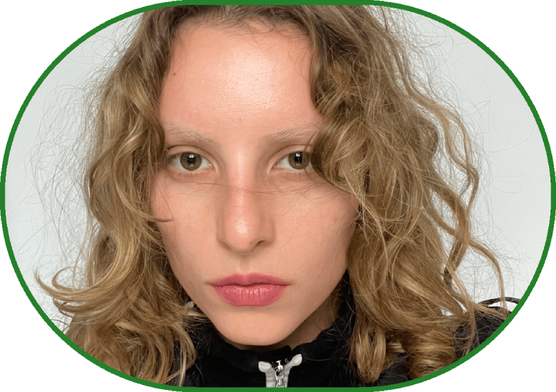 Close-up, portrait. A woman wearing black attire with a white zipper, with light brown eyes and long, curly light hair, looks intently at the camera against a white wall.