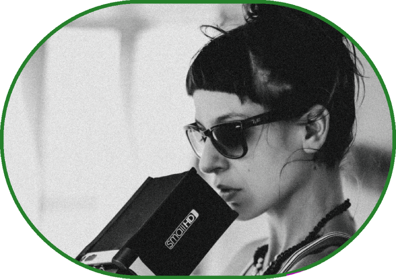 Великий план, чорно-білий портрет. Жінка в сонячних окулярах, з довгим темним волоссям, зібраним у пучок на потилиці, у намисті й легкій смугастій майці зосереджено дивиться в накамерний монітор.