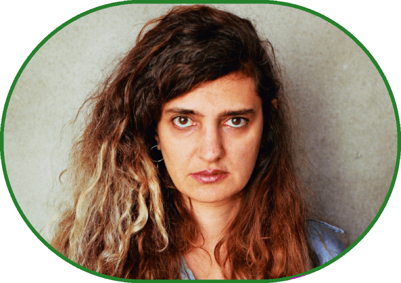 Medium shot, portrait. A woman stands facing forward against the backdrop of a greenish wall, wearing jeans and a blue long-sleeved shirt. She has long, loose, curly brown hair with a yellowish patch; her dark eyes gaze directly into the camera.