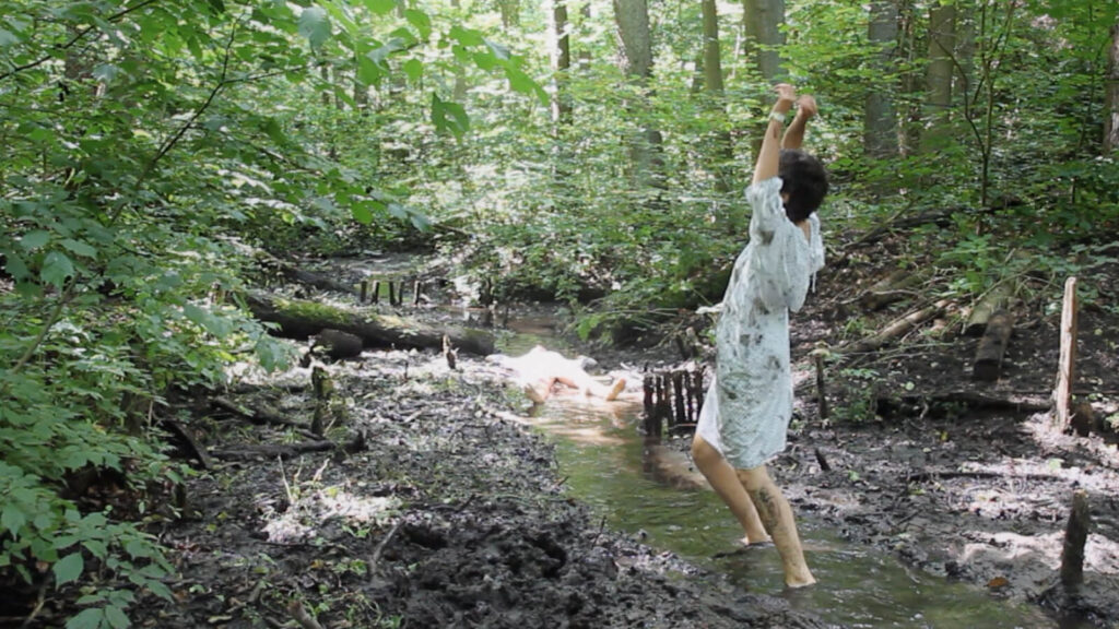 Wide shot. Gentle light streams through tree branches. A lively little stream runs through the lower part of the frame. Closer to the camera, a person in a hospital gown dances barefoot in the water. A bit farther, a person lies on their back with arms and legs spread out. Their silhouette fades in the bright rays of light.