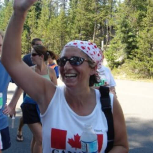 Medium close-up, portrait. A woman wearing sunglasses and a white bandana patterned with red maple leaves is running, smiling joyfully, and waving. She is wearing a white tank top with a Canadian flag, and holding a bottle of water in her hand. Behind her are other runners and trees in bright sunlight.